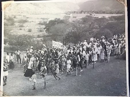 A black and white photograph of a masquerade band, stilt walkers among them, leading a large crowd across open ground below the hills.